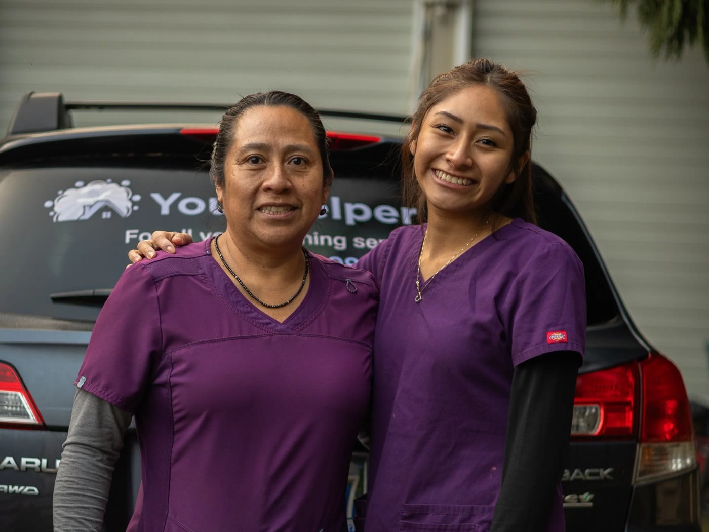 Celiflora and her daughter standing with their arms around each other, both in purple scrubs, smiling straight at the camera. Behind them is a dark Subaru Outback with a white decal across the rear window reading Your Helper, for all your cleaning services.