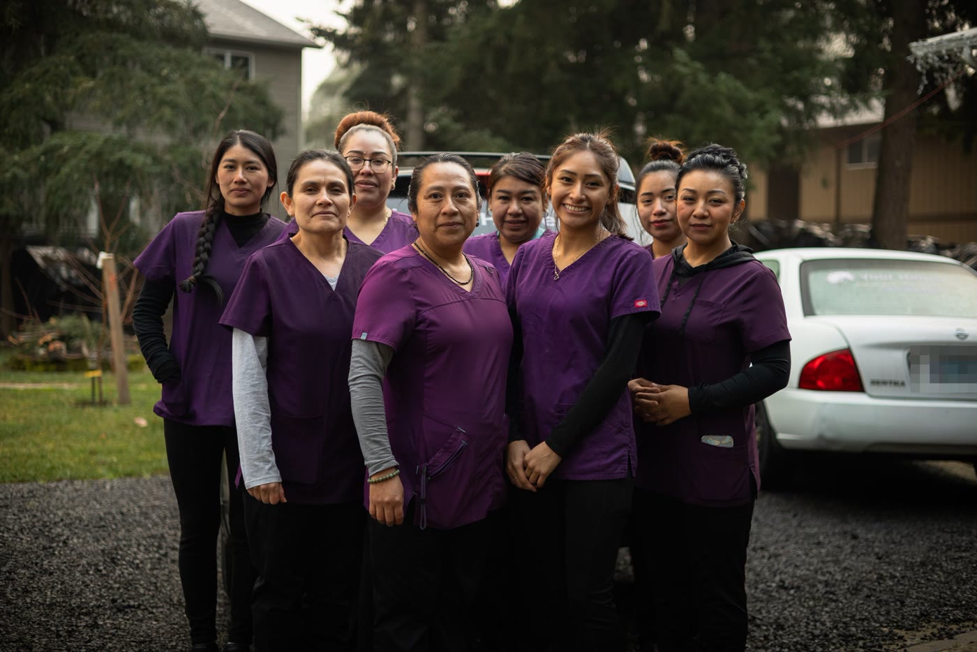 Eight women of the Your Helper team standing together in a gravel driveway, all in purple scrubs, with Celiflora in the centre. Misty trees rise behind them, and a white Nissan Sentra at the right of the frame carries a Your Helper decal in its rear window.